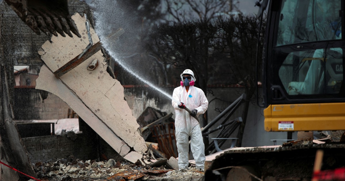 A person in a white suit and a protective face mask is using a hose to spray a stream of water.
