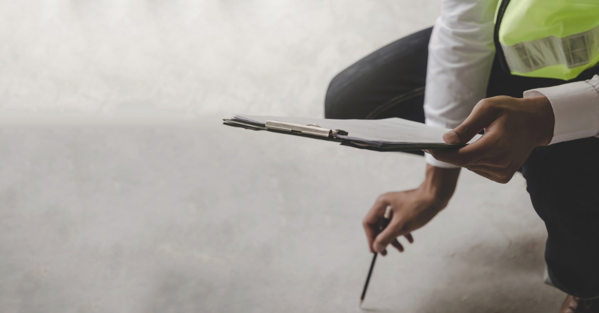 A man is holding a clipboard in one hand and a pencil in the other. He’s looking at the clipboard while kneeling.