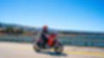 A motorcycle rider on a red-and-black adventure motorcycle driving down a road with a lake background.