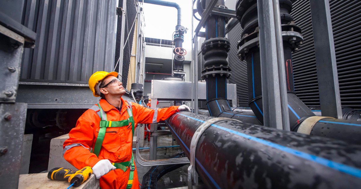 A man dressed in orange is looking at several pipes; he has his hand resting on one of the pipes.