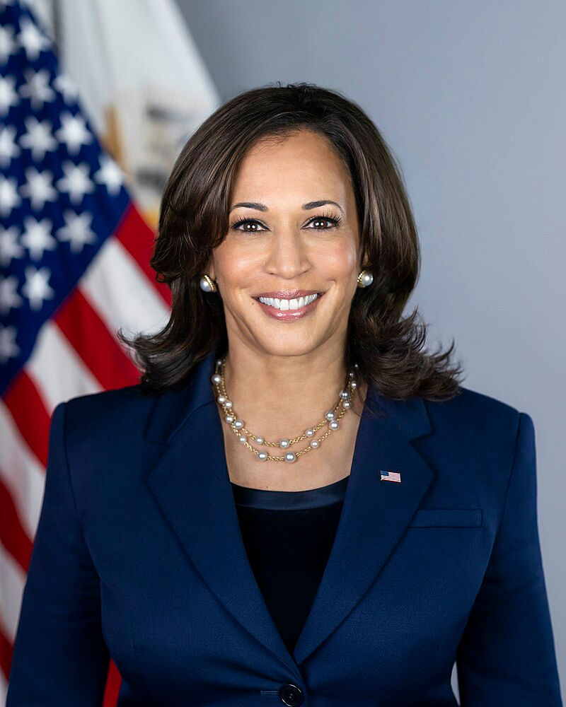 Woman in a navy suit smiles against a gray backdrop. An American flag is partly visible.