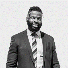 Smiling man in a suit with a striped tie stands confidently against a plain background. The image is black and white.