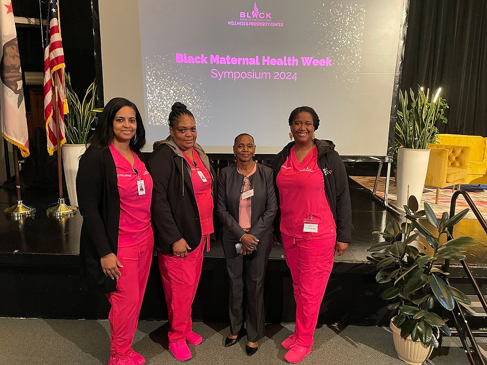 Four women pose on stage at the Black Maternal Health Week Symposium 2024. Three wear pink scrubs. Text on screen; flags and plants in view.