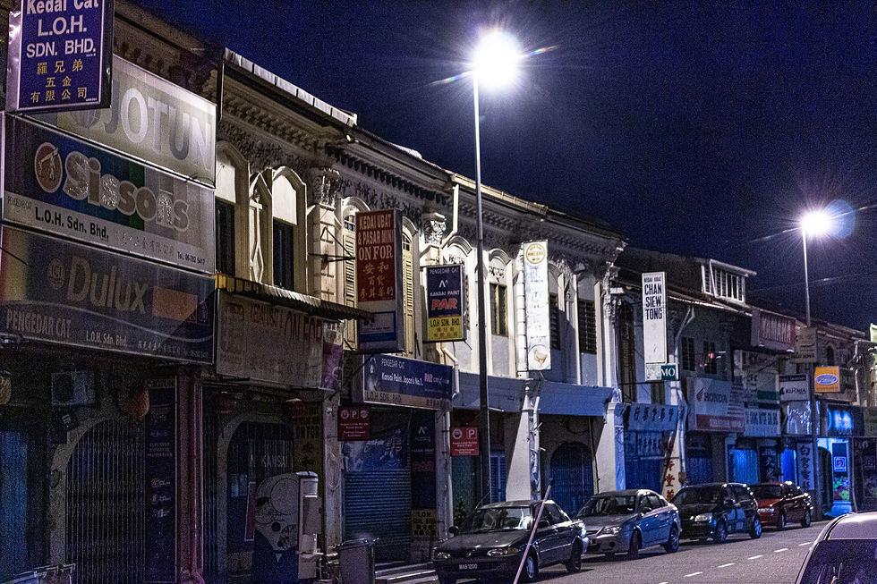 Night view of a street with old buildings and bright streetlights. Various shop signs in different languages are visible. Cars line the street.