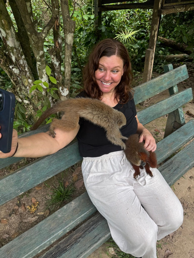 Woman on a bench in forest, smiling while taking a selfie with two small monkeys on her lap, one climbing her arm. Wearing black and white.