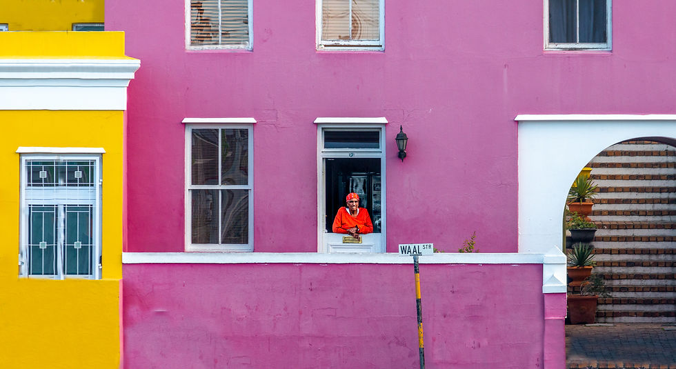 Person in orange, red headscarf leans from pink house window. Yellow adjacent building. Waal Street sign. Bright, lively atmosphere. Bo-Kaap