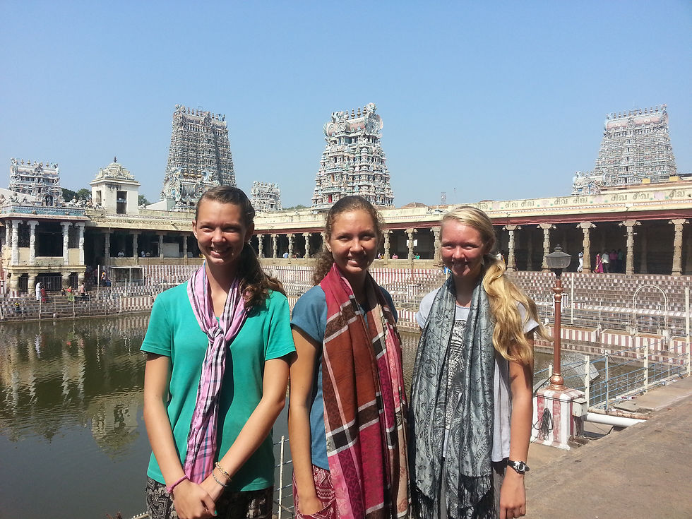 Three smiling people in colorful scarves stand before a historic temple with intricately carved towers. A water body and clear blue sky in the backdrop.