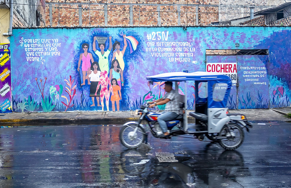 A mural with women on a blue wall, text advocating against violence. A person rides a motorcycle taxi on a wet street.