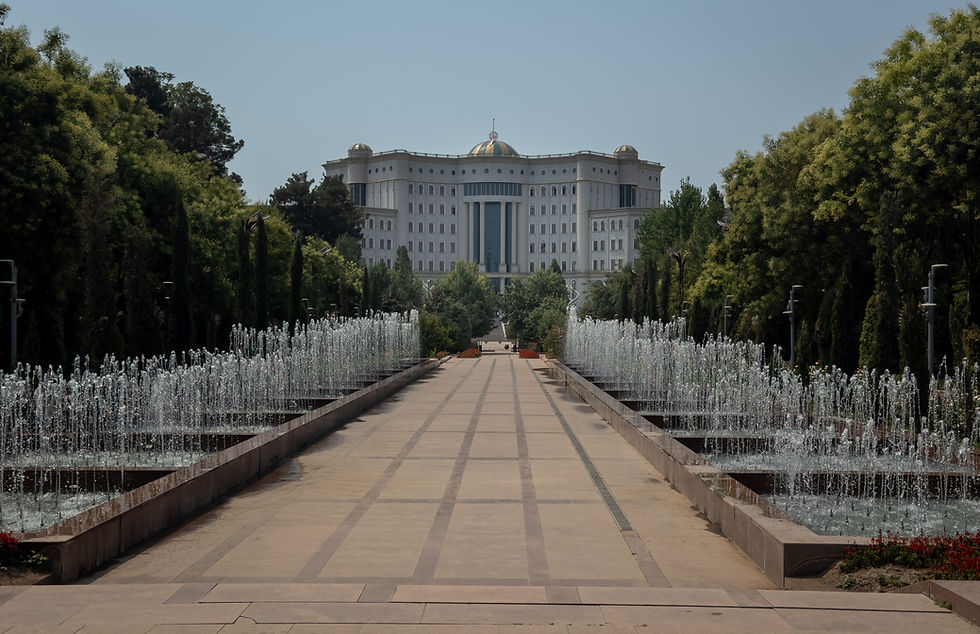 Path lined with water fountains leads to a grand white building with domes. Surrounded by lush trees under a clear blue sky.