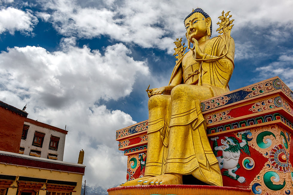 Golden Buddha statue on ornate pedestal under a blue sky with clouds at Likir Gompa. Vibrant patterns and nearby traditional building enhance serenity.