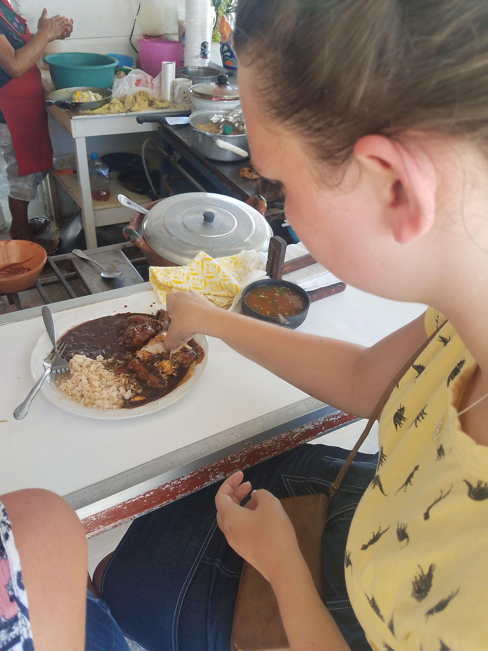 Woman in yellow shirt eats rice and sauce with chicken at a counter. Background shows a cook in a kitchen with pots and food prep.