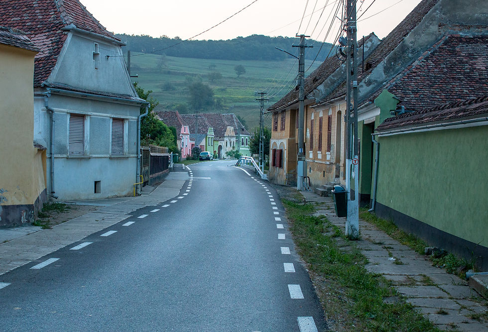 Narrow winding street with colorful houses, green hills in the background, and a parked car. The mood is calm and serene.