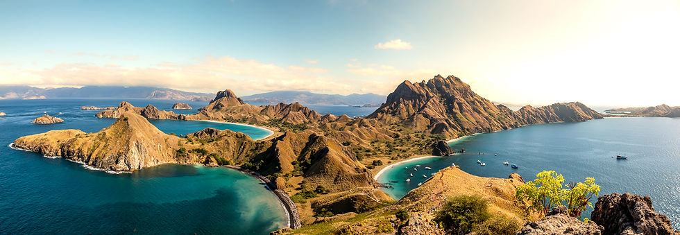 Sunlit island landscape with rugged hills and turquoise water. Boats dot the sea. Clear sky and distant land create a serene atmosphere.