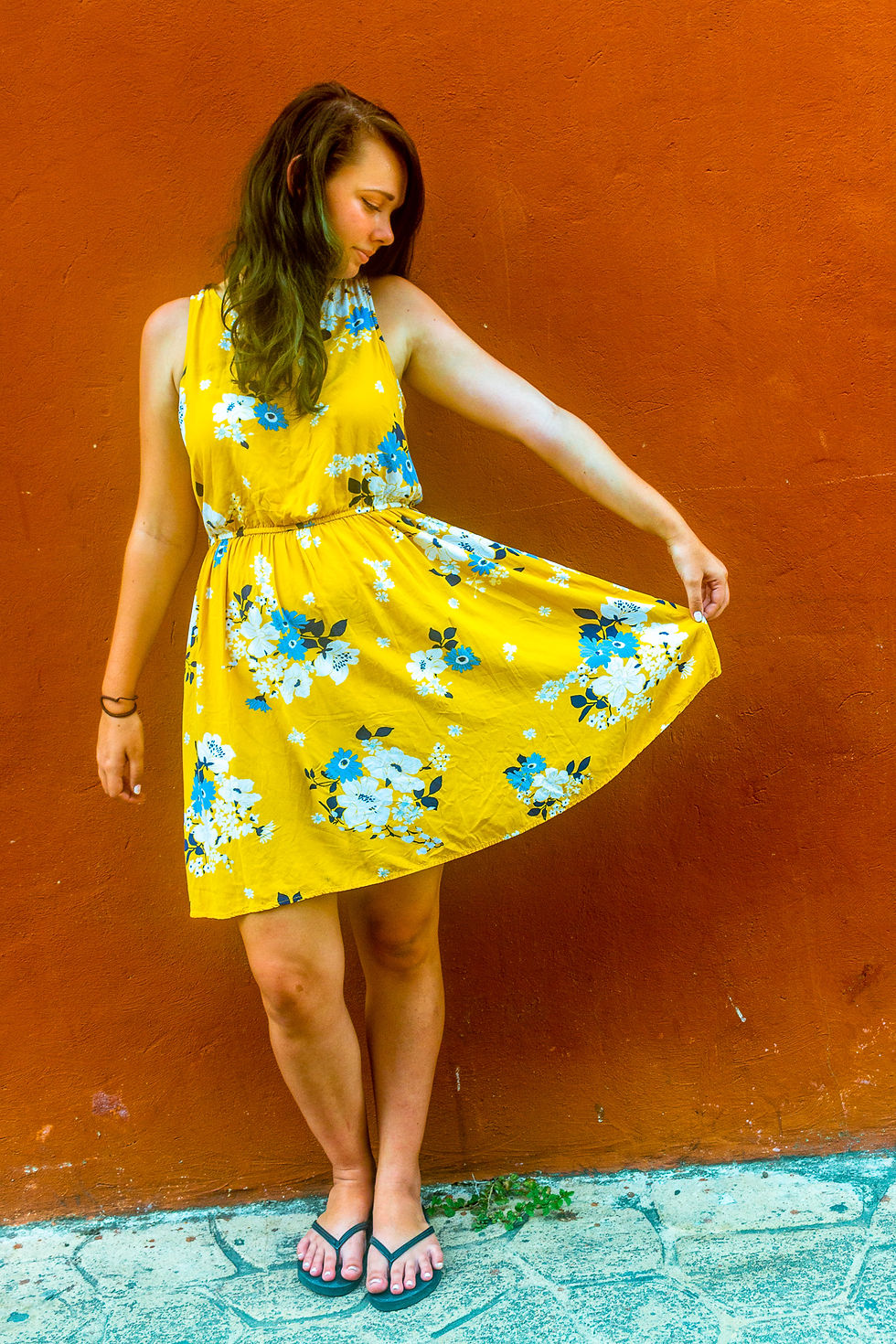 Woman in a yellow floral dress poses against an orange wall, gently holding the skirt. She wears flip-flops on a stone pavement.