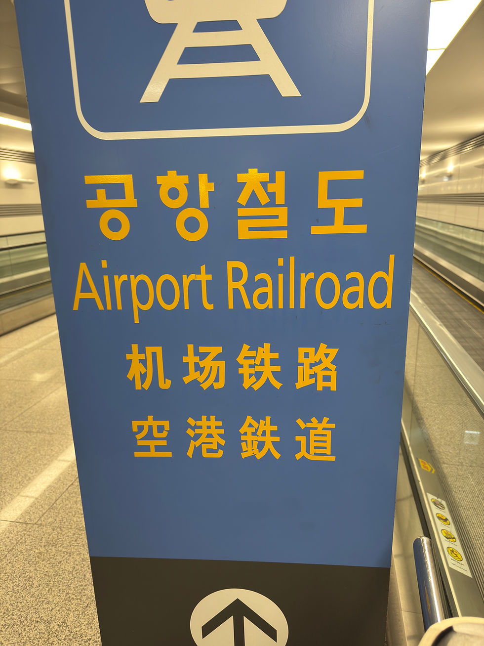Blue sign with yellow text in Korean, English, and Chinese for "Airport Railroad" near a moving walkway in a modern, well-lit station.