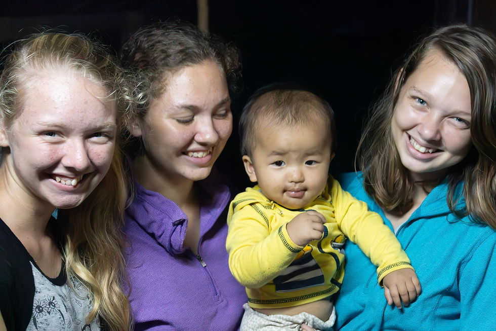 Three women smiling and a baby in a yellow shirt looking curious. Dark background, casual attire, joyful atmosphere.