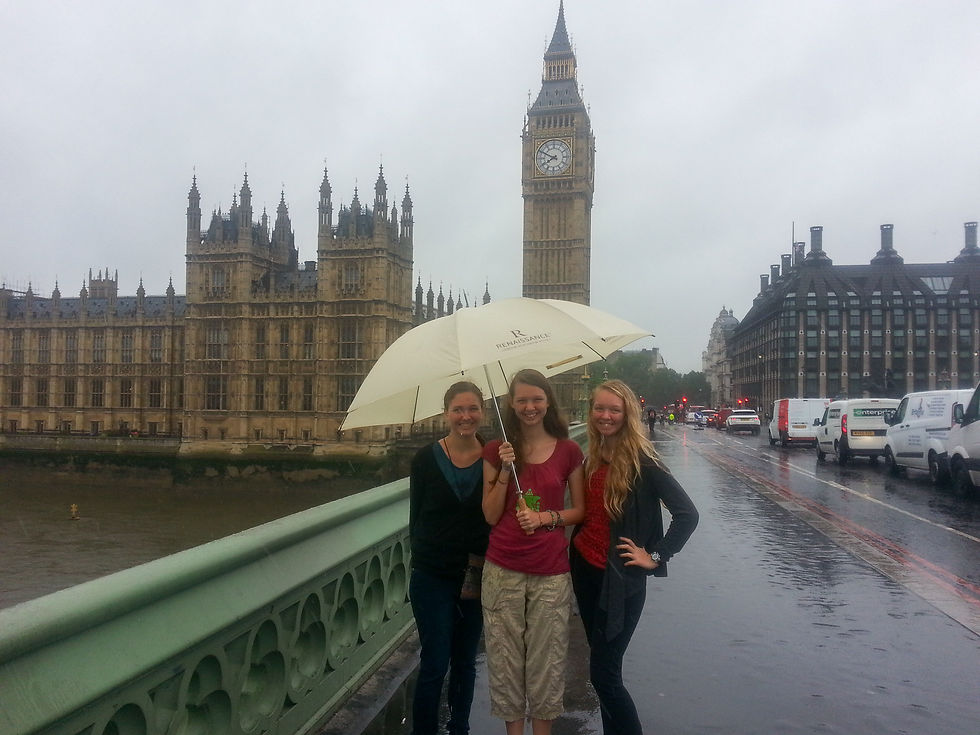 Three people smiling under an umbrella on a rainy day with Big Ben in the background. Overcast sky, wet pavement, and passing vehicles.