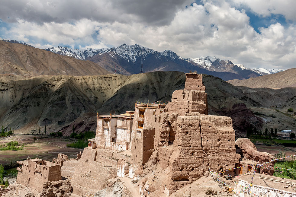 Bagso Gompa with prayer flags on a rocky hill against snow-capped mountains and cloudy sky. Brown and white tones dominate the scene.