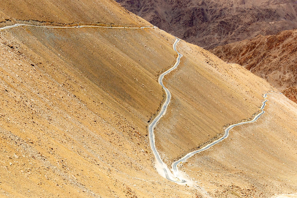 Winding road on a barren, rocky mountain slope with a desolate, rugged landscape. Browns and beiges dominate the scene. No visible text.