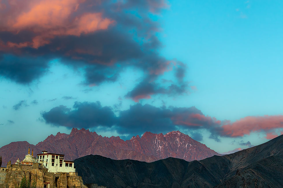 Lamayuru Monastery perched on rocky hill against backdrop of jagged mountains. Colorful sunset sky with pink and blue clouds creates a serene mood.