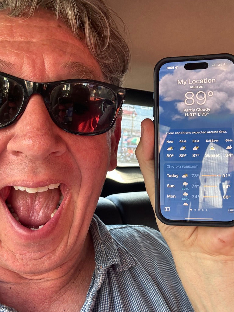 Man wearing sunglasses happily shows a phone displaying weather info for Iquitos, 89°F, partly cloudy. Interior car setting.
