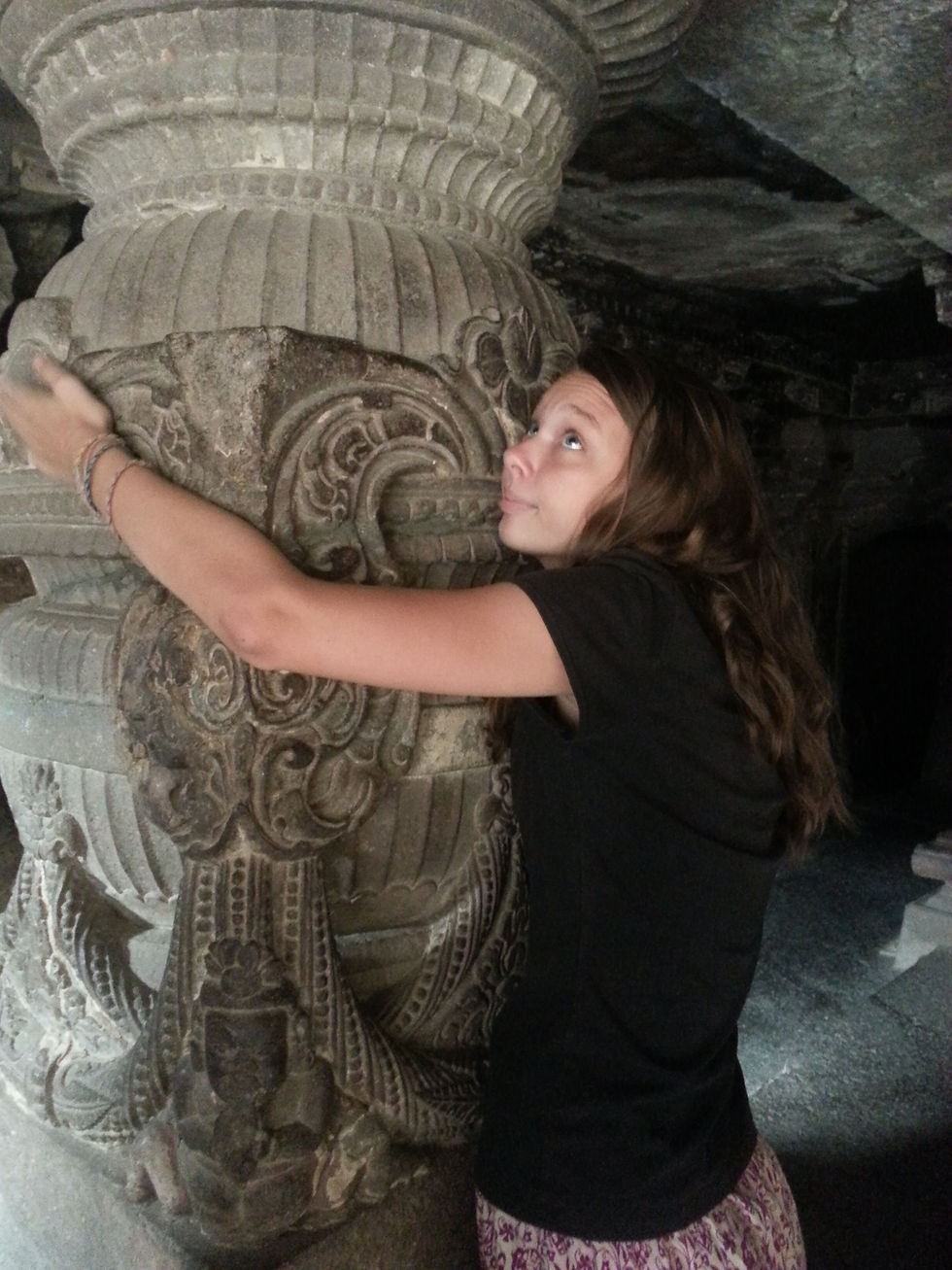 Woman hugging an intricately carved stone pillar in a dimly lit ancient setting, wearing a black shirt and patterned skirt, looking content.