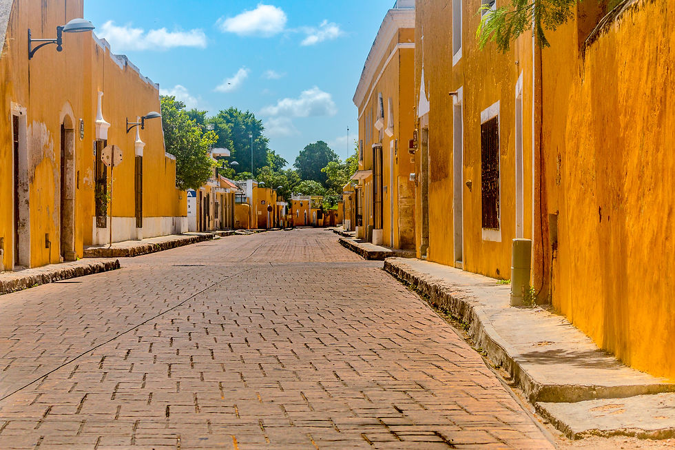 Quiet cobblestone street lined with bright yellow buildings under a blue sky with fluffy clouds. Trees peek above rooftops. Izamal
