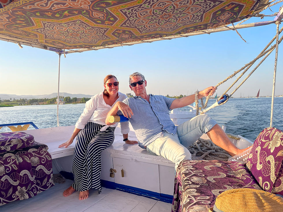 A couple relaxes on a boat with a patterned canopy. The woman wears a white shirt and striped skirt; the man holds a rope. Calm river backdrop.