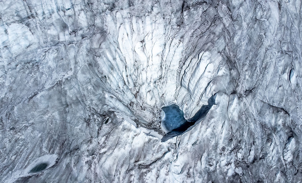 Aerial view of a glacier with white and gray ice patterns. A small dark blue meltwater lake appears near the center, showcasing icy textures.