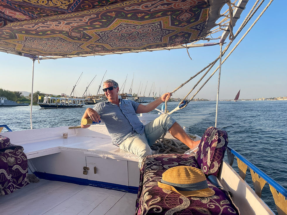 Man relaxing on a patterned boat with a calm river and sailboats in the background. Wears sunglasses, hint of a smile. Hat on bench.