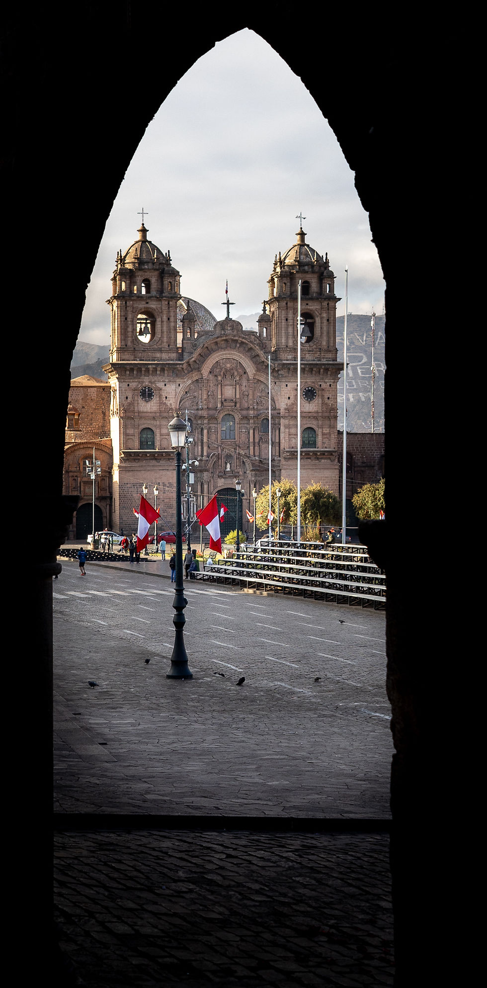 Arched view of a historic cathedral with twin towers in the background. Peruvian flags line a cobblestone street, adding vibrant reds.