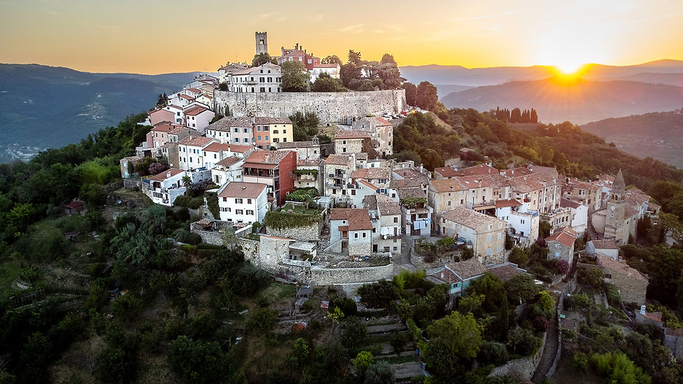 Hilltop village at sunset with stone houses and red roofs, surrounded by lush greenery and mountains. Sun casts a warm glow over the scene.