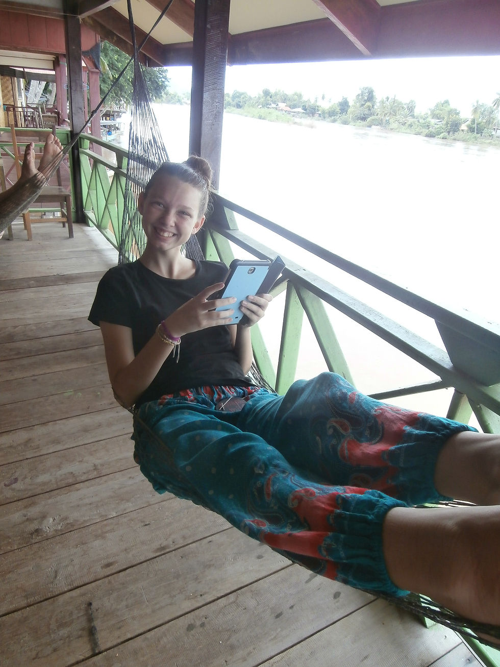 Girl in a black shirt and colorful pants relaxes in a hammock, holding a phone, smiling on a wooden porch overlooking a river.