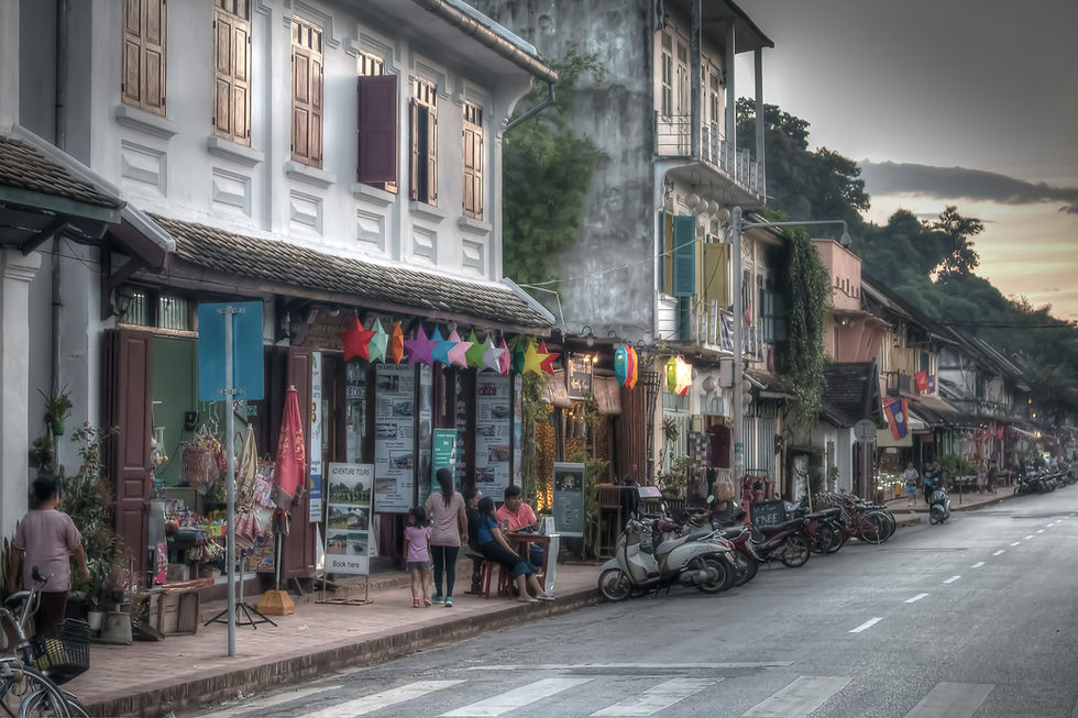 Street view with colorful paper stars hanging above shops. People walk, sit, and interact near bikes. Warm, inviting evening ambiance.