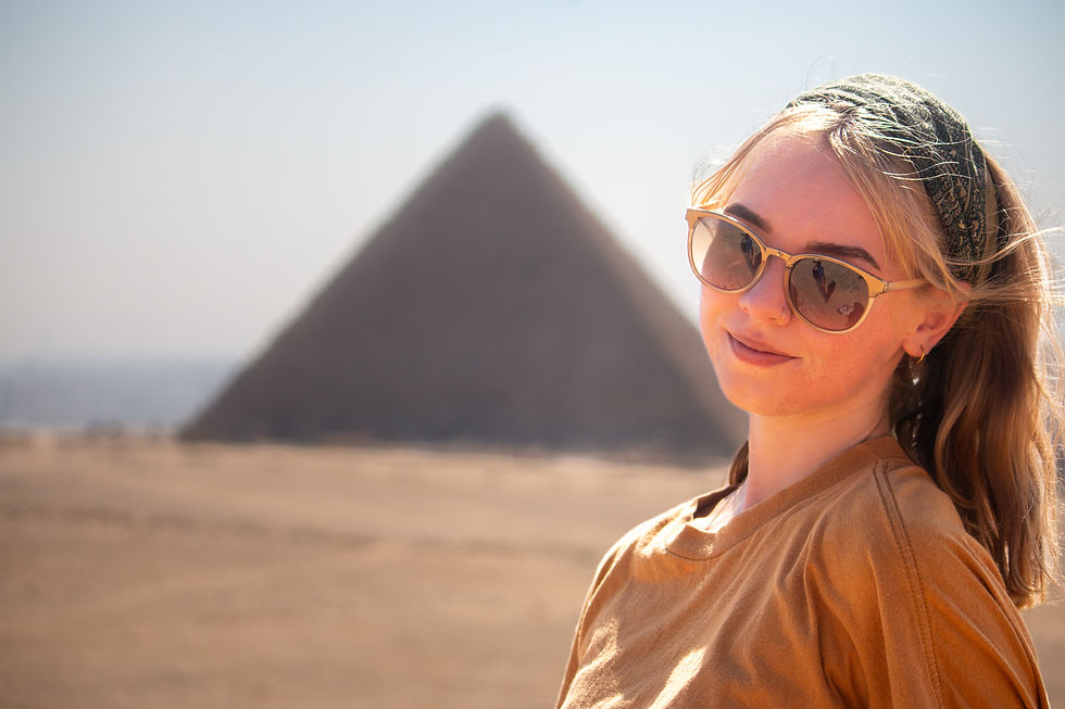 Woman in sunglasses smiling with a pyramid in the background. She wears a headband and a brown top. Bright, sunny desert setting.