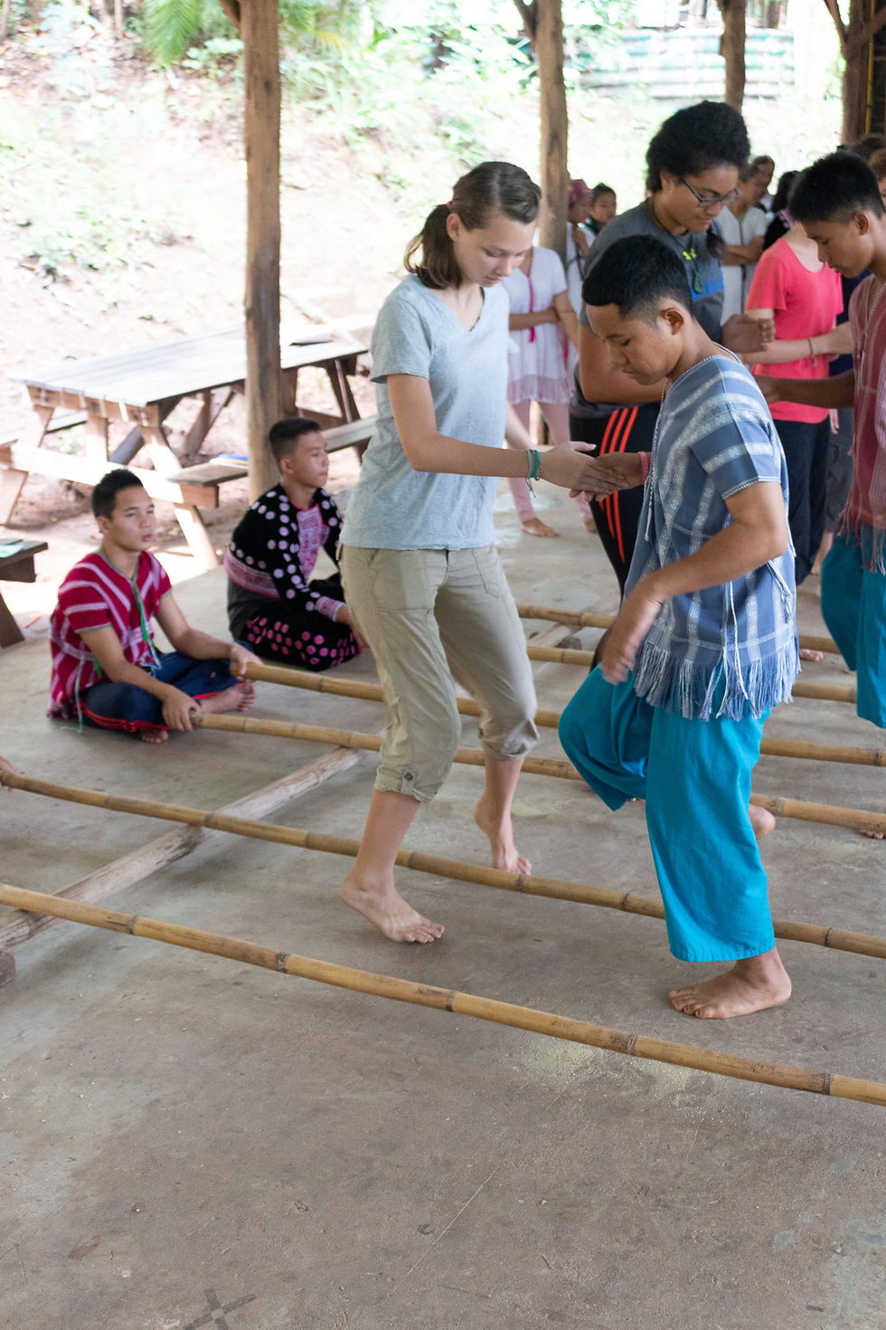 People dance between bamboo poles in an outdoor setting. They're barefoot, wearing colorful clothes, surrounded by seated onlookers.