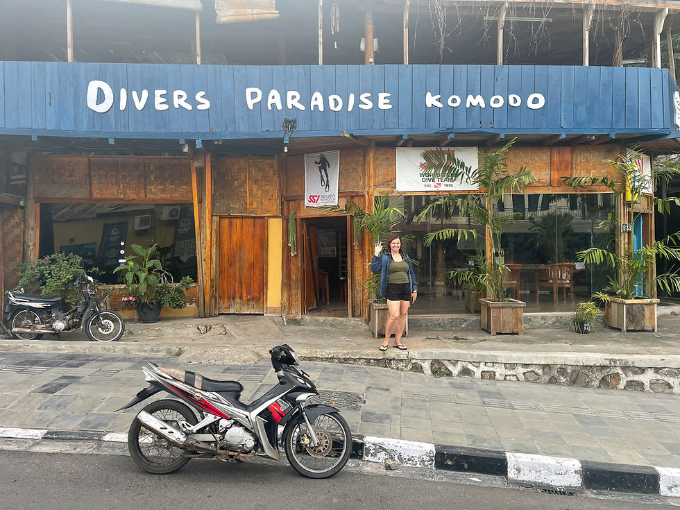 Person waving outside "Divers Paradise Komodo" with a blue sign. A motorcycle is parked in front. The mood is casual and welcoming.