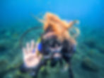 Scuba diver with orange hair waves underwater with rocks and plants in the background. Wearing gear with "Cressi" visible. Calm mood.