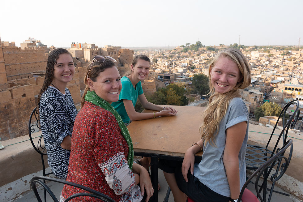 Four people smiling at a rooftop table in a desert town, surrounded by brown buildings. Casual attire, warm ambiance.