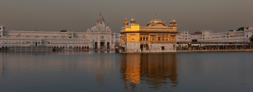 Golden temple glowing at sunset, surrounded by a calm water body and white architecture. Reflection visible, peaceful ambiance.