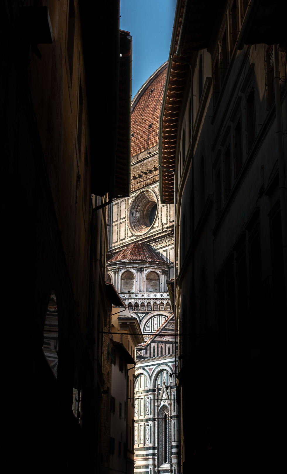 Florence Cathedral dome framed by dark alley shadows; sunlit marble facade with intricate patterns; vivid blue sky above. Tranquil setting.
