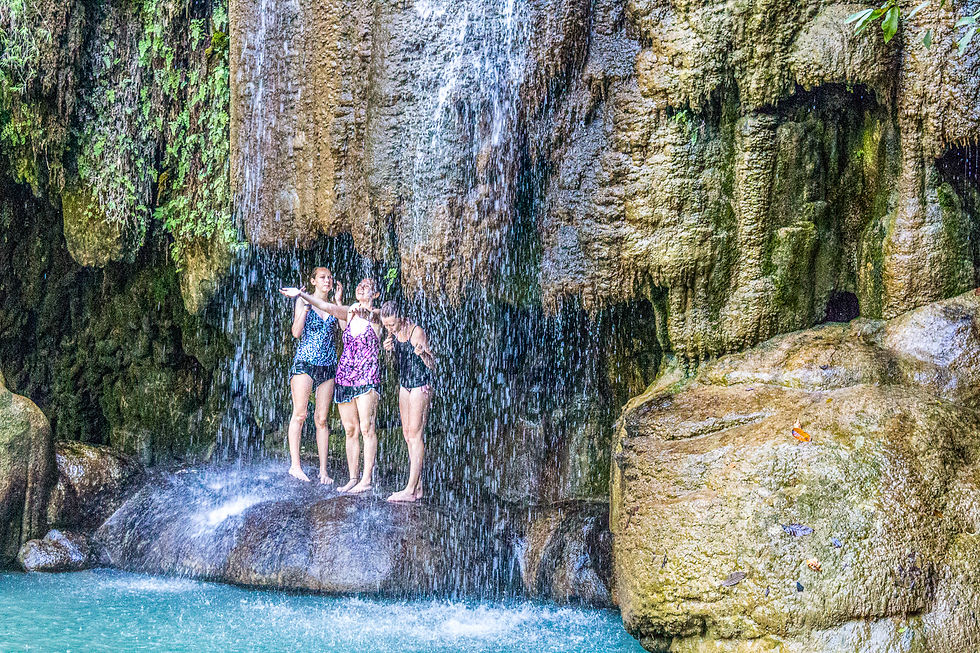 Three people stand under a waterfall, enjoying the water. They're on a rocky ledge with lush greenery. The mood is joyful and refreshing.