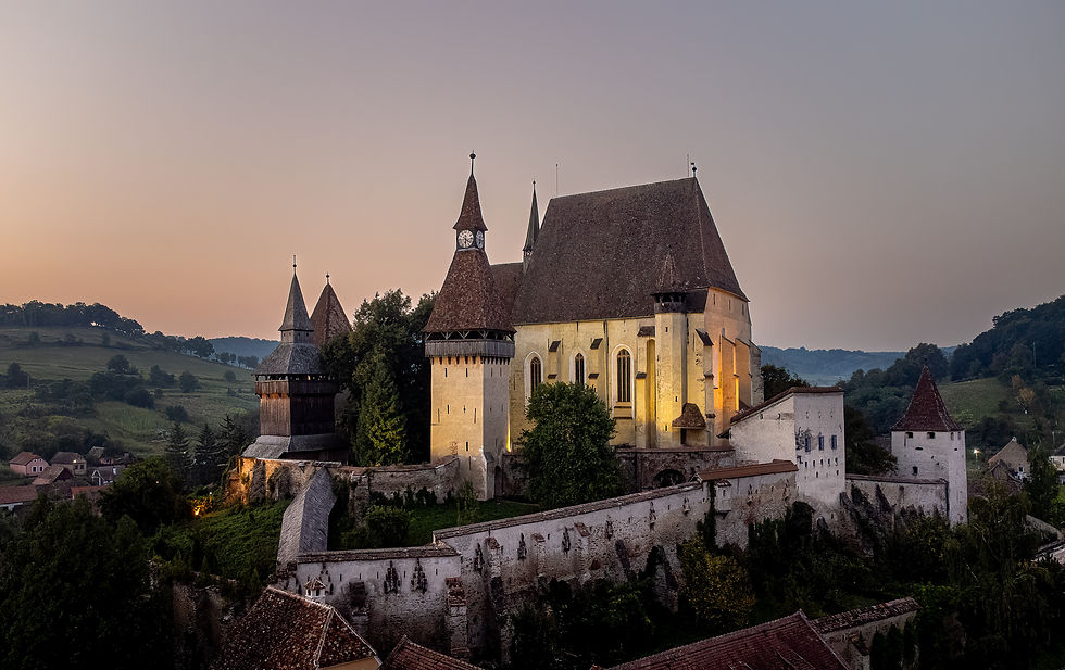 Historic stone church with tall towers at sunset, surrounded by medieval walls and lush greenery. A peaceful village lies in the background.