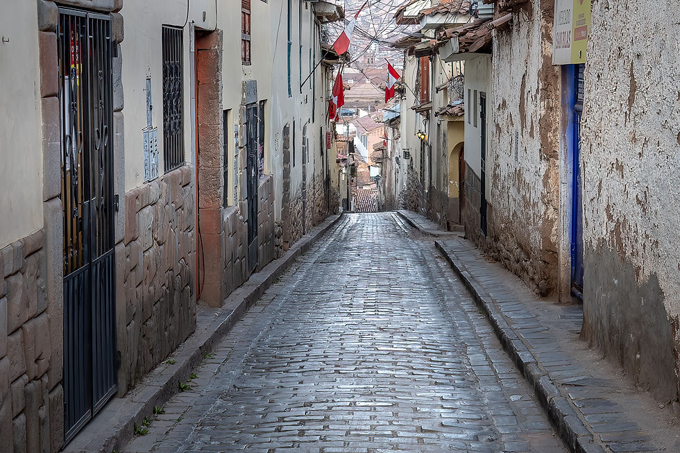 Narrow cobblestone street with weathered buildings, red and white flags on walls, and a distant hilly backdrop. Calm and picturesque.