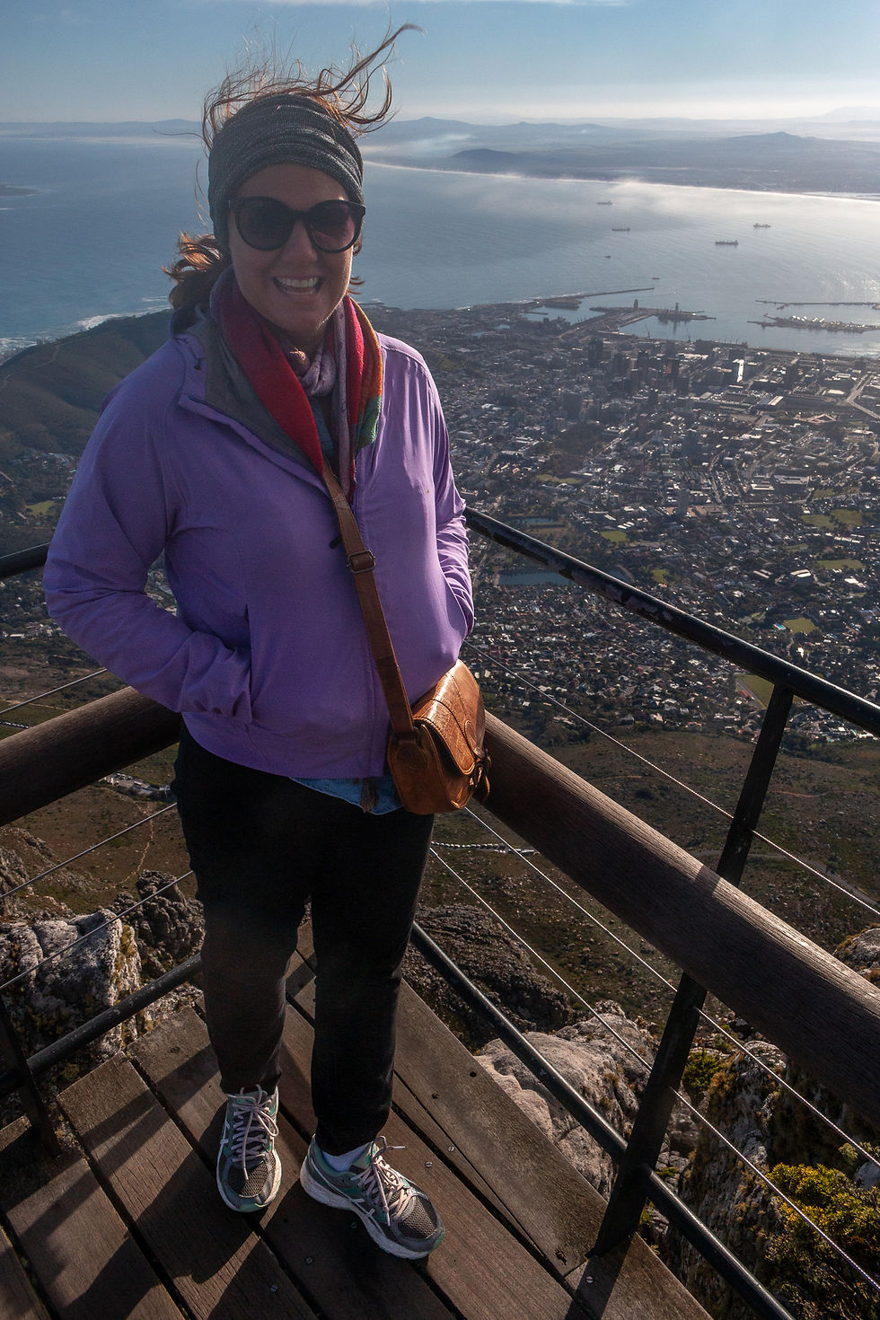 Smiling person in a purple jacket stands on a wooden platform overlooking a coastal cityscape, with windy hair and a crossbody bag.