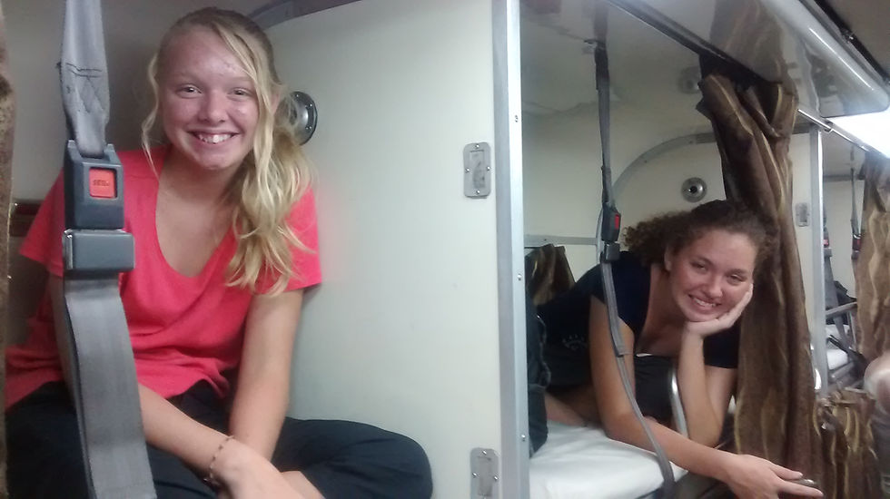 Two smiling women in an overnight train berth, one in a pink shirt and the other leaning on an elbow. Relaxed mood, beige curtains.
