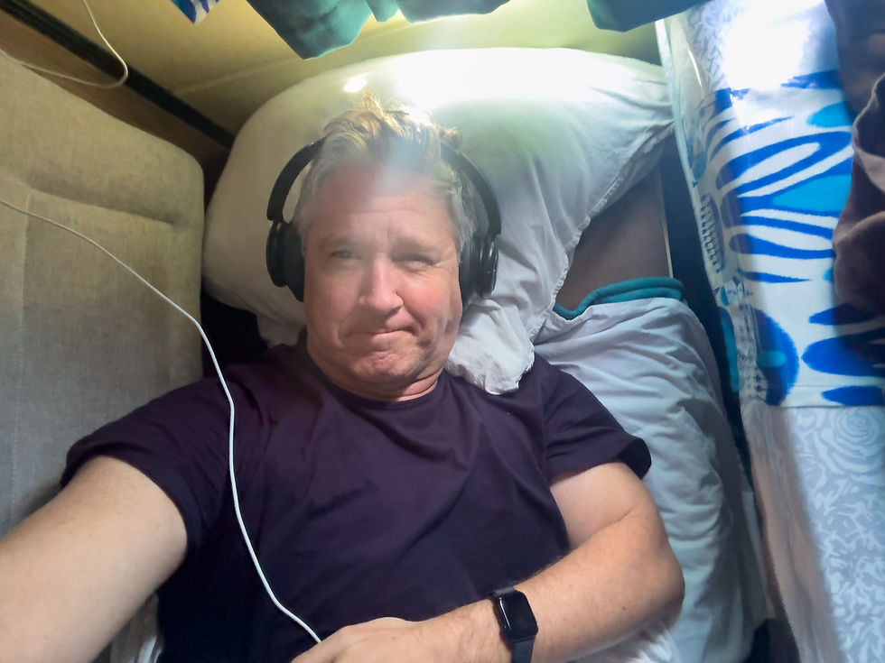 Man lying on bed, wearing headphones and a dark shirt. Sunlight shines on his face. Blue patterned curtain in background. Relaxed mood.