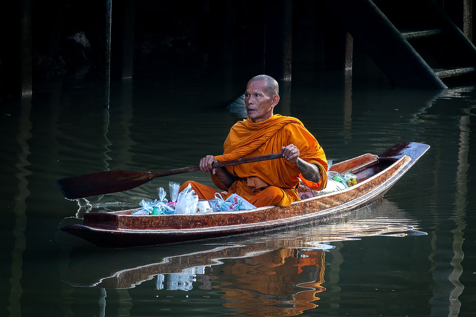 Monk in orange robe paddles small wooden boat on calm water, bags of items aboard, shadowy background, serene mood.