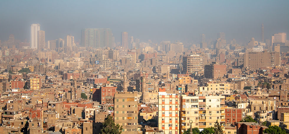 Cityscape of a densely packed urban area with beige, red, and orange buildings under a hazy blue sky. Tall structures in the distance.