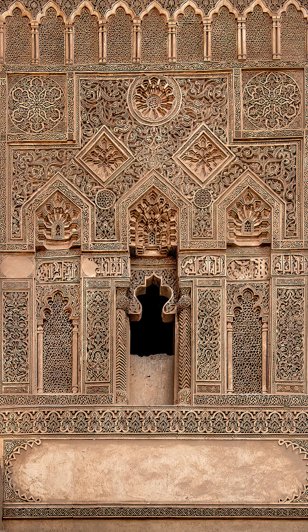 Intricately carved stone wall with geometric and floral patterns, Arabic script, and ornate arches. Beige tones, historical architecture.